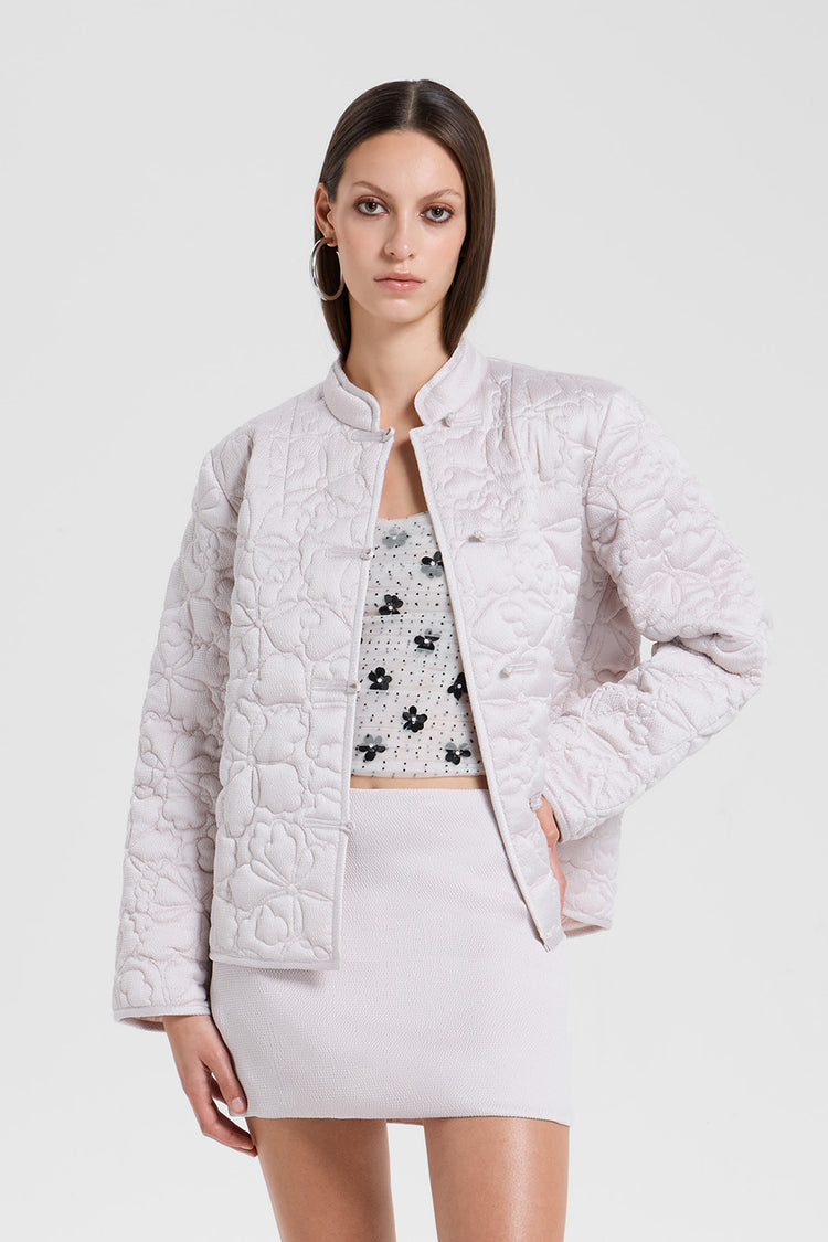 Woman wearing a light gray quilted jacket over a floral top and white skirt on a white background
